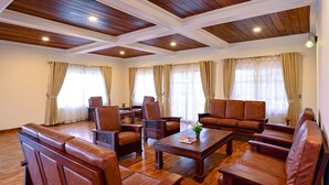 Un salón comedor amplio con suelo de madera, techo alto con vigas de madera y ventanas grandes con cortinas color beige.