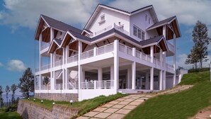A large, multi-story house with a prominent balcony, situated on a grassy hill with a clear sky. - Petals Luxury Homestay (Kunda)