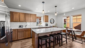 A modern kitchen with wooden cabinets, a stainless steel refrigerator, a large island with stools, and pendant lighting. - The Bear's Den - Dover ID (Dover)