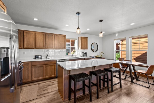 A modern kitchen with wooden cabinets, a stainless steel refrigerator, a large island with stools, and pendant lighting. - The Bear's Den - Dover ID (Dover)