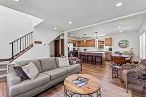 A spacious living area with a sofa, coffee table, and wicker chairs. A kitchen with wooden cabinets and a central island is visible in the background. - The Bear's Den - Dover ID (Dover)