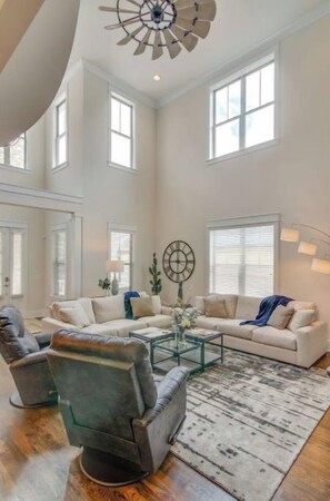 A spacious living room with a high ceiling, large windows, and a central coffee table.