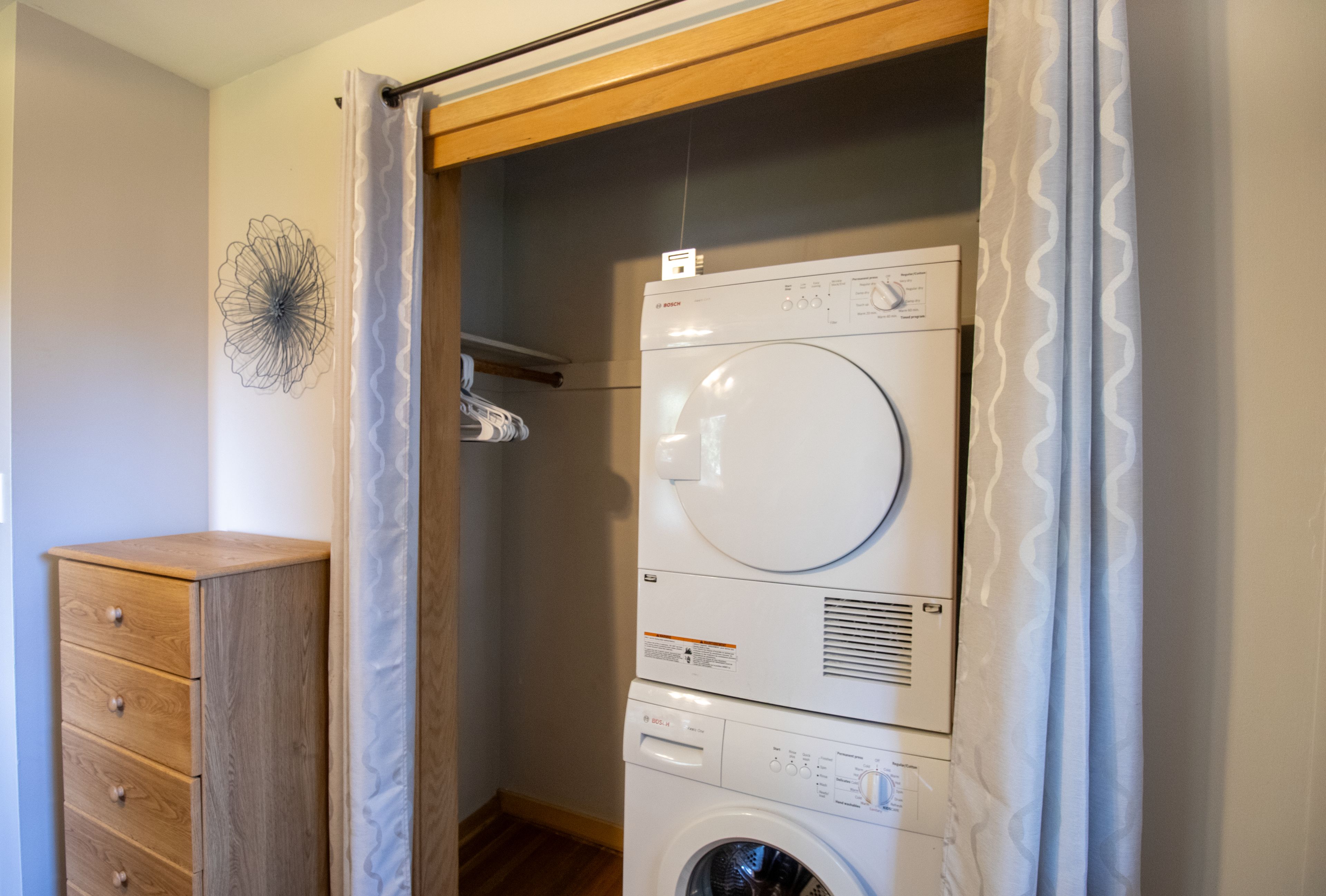 Une petite buanderie équipée d’un lave-linge et d’un sèche-linge superposés, d’un meuble en bois et d’un élément décoratif mural.