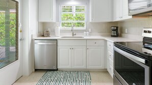 Une cuisine compacte avec des meubles blancs, un lave-vaisselle encastré, une fenêtre avec des stores verts, une machine à café et un four à micro-ondes.