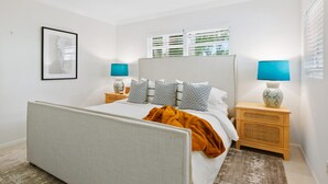 Une chambre à coucher comprenant un lit, deux tables de chevet, une commode, une fenêtre avec des stores, un tableau encadré au mur et une lampe sur pied.