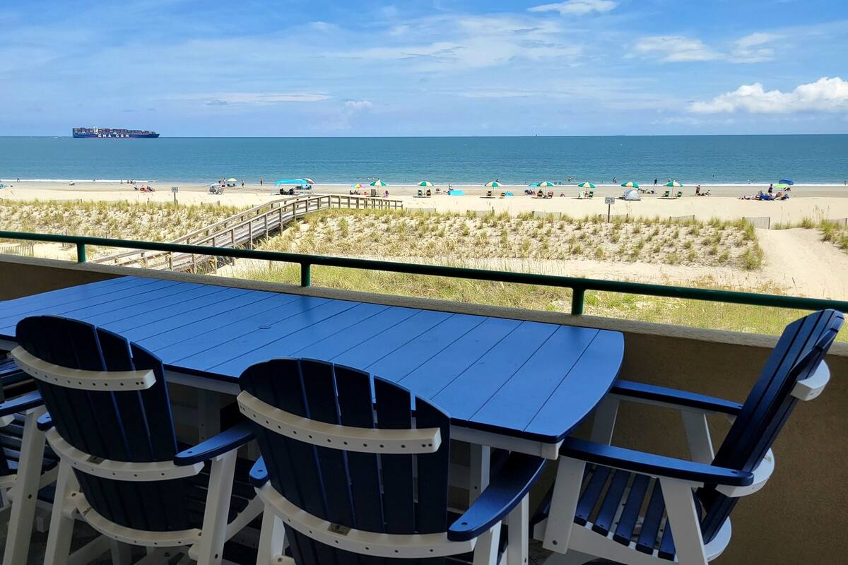 A terrace with a table and chairs overlooking a beach with umbrellas and a ship in the distance.