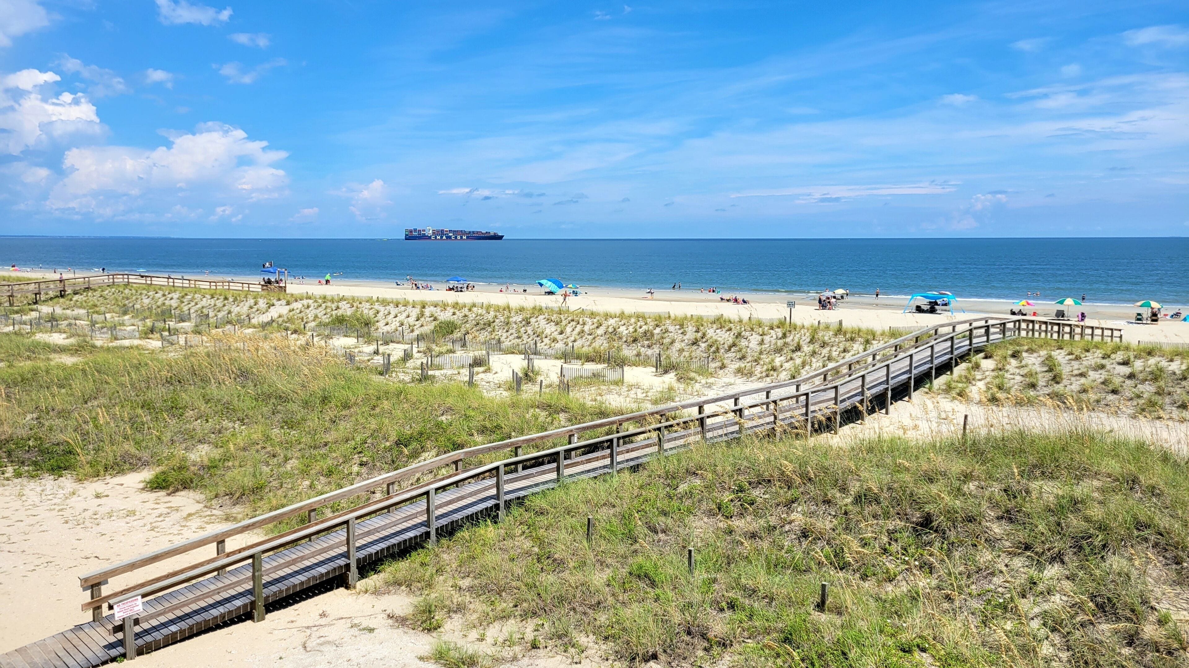 Une plage avec une promenade en bois, des dunes de sable et un navire à l’horizon.