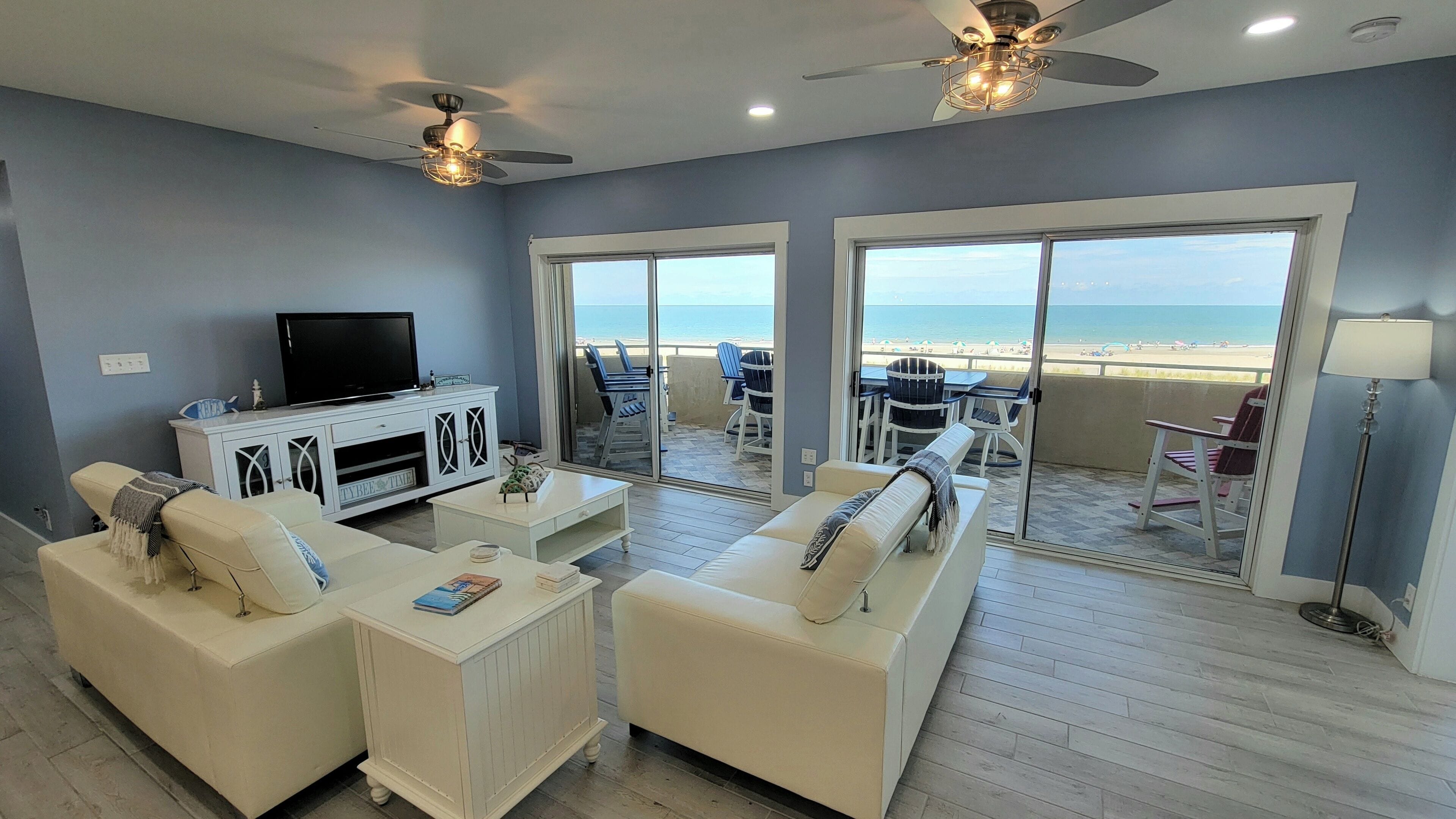 Un salón con televisor, sofás blancos y un balcón con vistas a la playa.