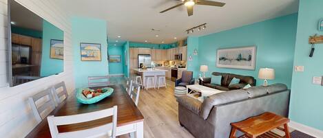 A modern kitchen with a central island, a dining area with a wooden table and white chairs, a grey sofa, and a ceiling fan.
