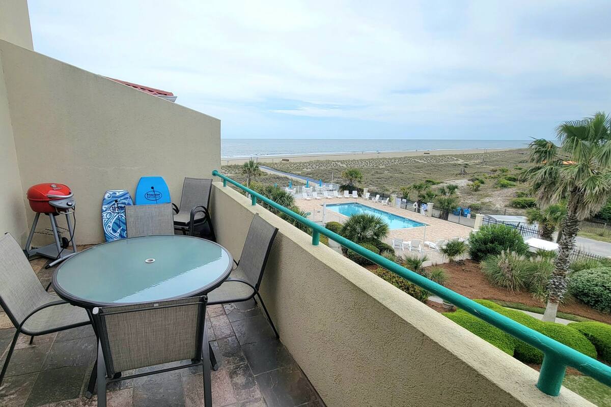 A balcony with a glass table and chairs, a barbecue grill, and two surfboards.