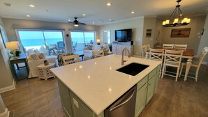 Un'ampia cucina con un'isola grande, una zona pranzo con sedie bianche e vista sull'oceano attraverso ampie finestre.
