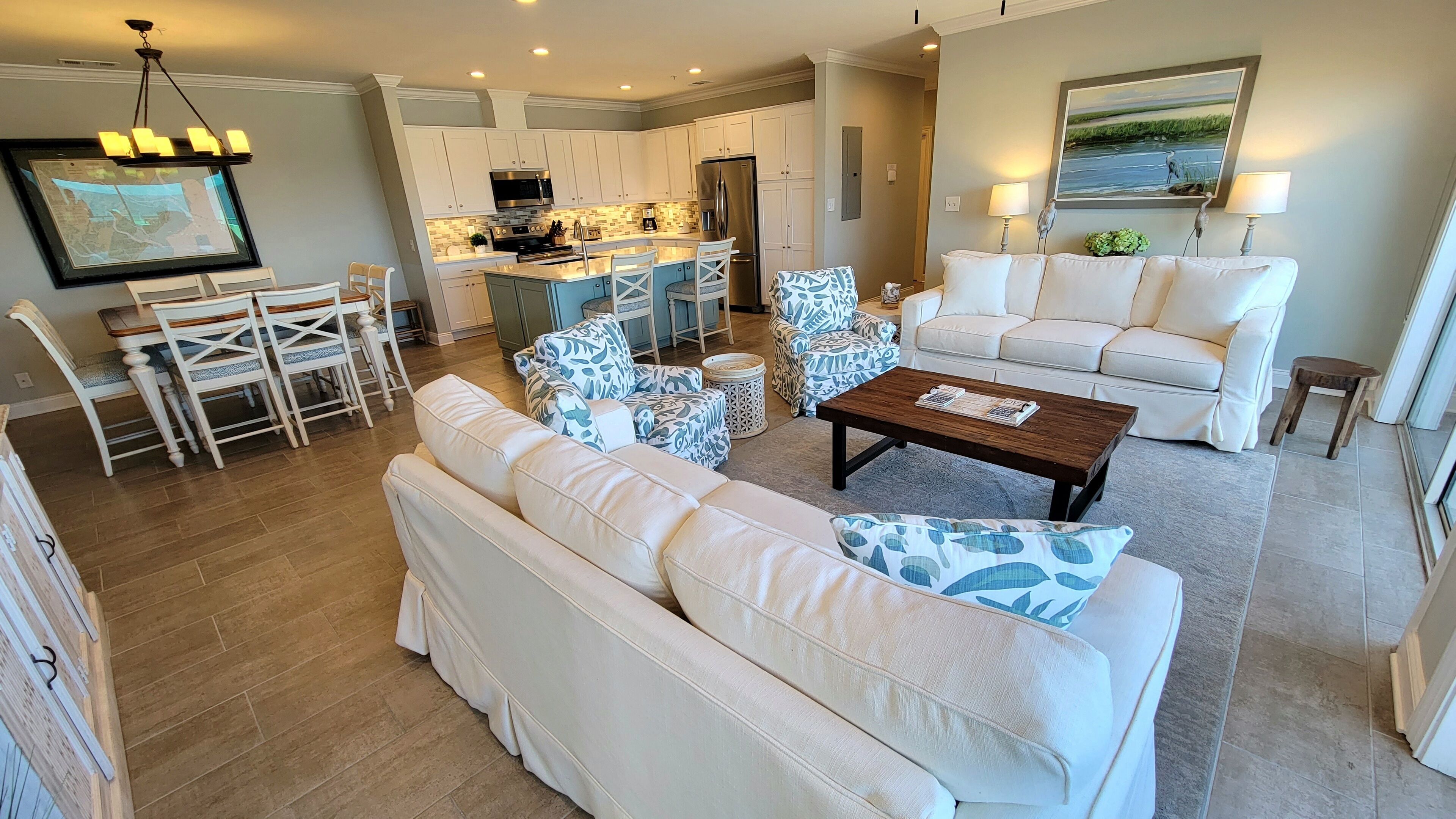 A spacious living room with a large white sofa, matching chairs, and a coffee table. A kitchen with white cabinets and modern appliances is visible in the background.