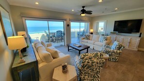 A spacious living room with a large window overlooking the beach, a ceiling fan, and a flat-screen TV mounted on the wall. - Desoto Beach Club 3 Tybee Vacation Rentals (Tybee Island)