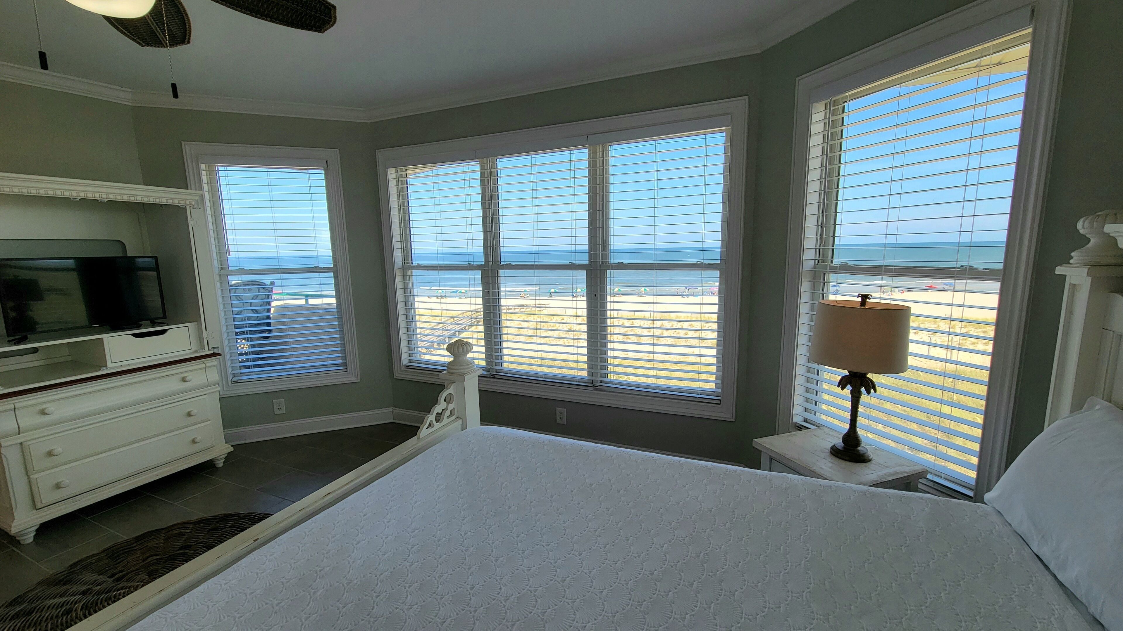 A bedroom with a bed, a dresser, a lamp, and large windows with beach views.