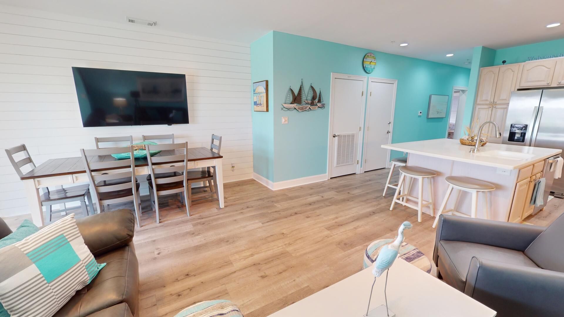 A modern living room with a wooden floor, a dining area with a table and chairs, a kitchen with a white countertop and bar stools, and a TV mounted on the wall.
