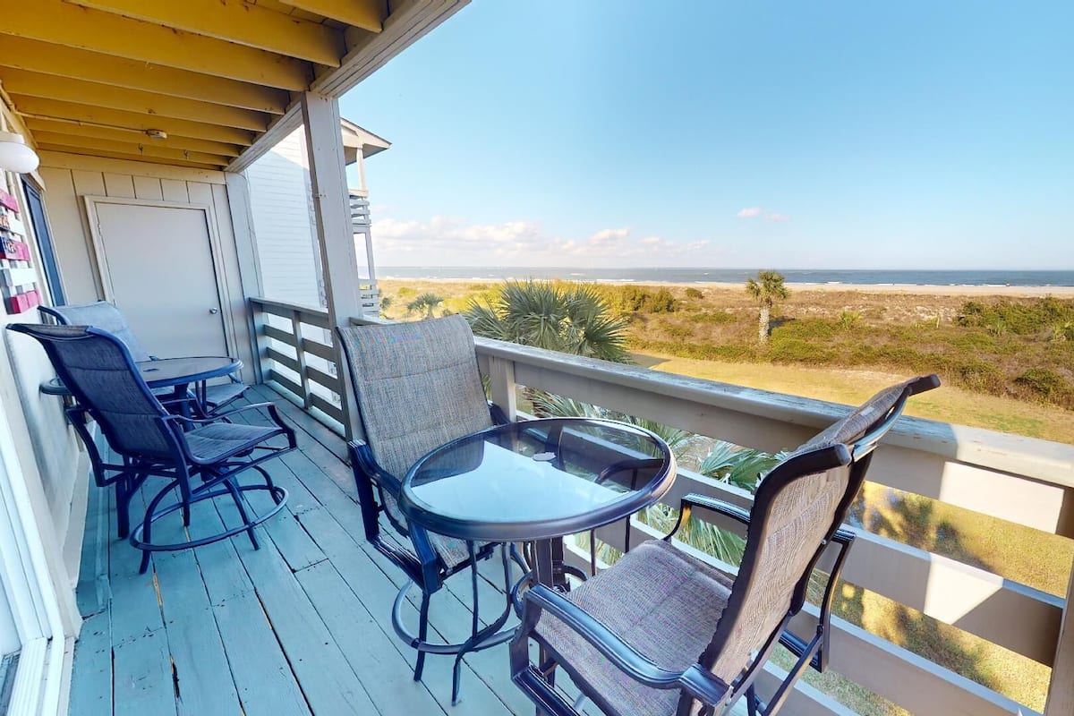 A balcony with a glass table and four chairs, overlooking a beach and ocean.