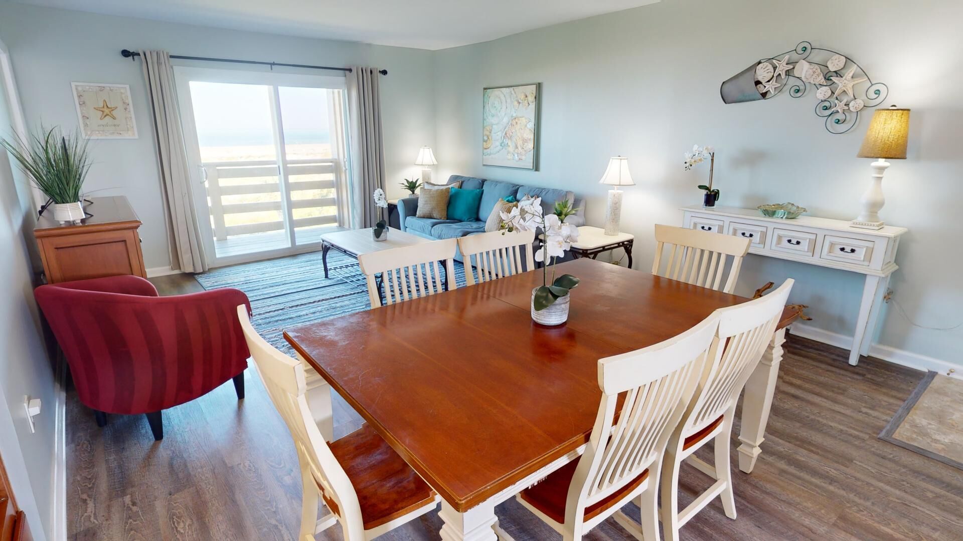 A dining area with a wooden table and white chairs, a sofa with blue cushions, and a view through a large window to the beach.