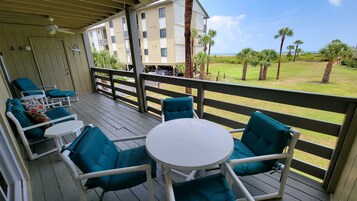 A covered outdoor seating area with chairs and a table, overlooking a grassy area with palm trees and a building in the background.