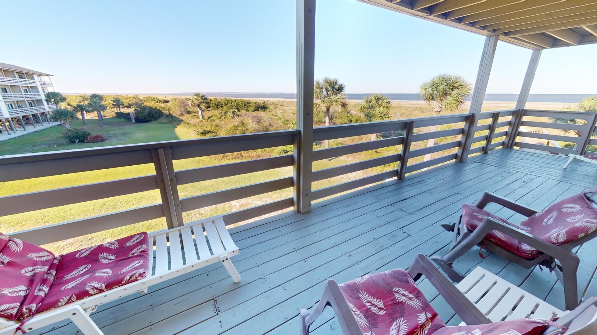A covered outdoor deck with white chairs and a table, overlooking a beach and a golf course.
