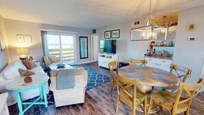 A spacious living area with a large window, a dining table with chairs, a sofa, and a television. - Lighthouse Point 3 by Tybee Vacation Rentals (Tybee Island)