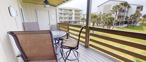 A balcony with a table and chairs, overlooking a pool area with palm trees and a building with balconies.