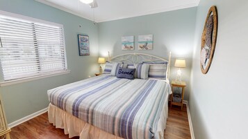 Une chambre à coucher comprenant un lit, deux tables de chevet, une fenêtre avec des stores et des œuvres d’art murales.