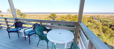 A covered outdoor area with a wooden deck, two wicker chairs, a small round table, and a view of the ocean and dry grassland.