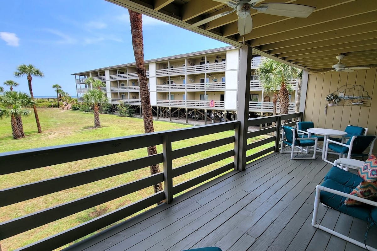 A wooden deck with outdoor furniture, a view of a building with multiple balconies, and palm trees.