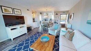 A modern living room with a TV, sofa, coffee table, and a kitchen area in the background. - Lighthouse Point 3 by Tybee Vacation Rentals (Tybee Island)