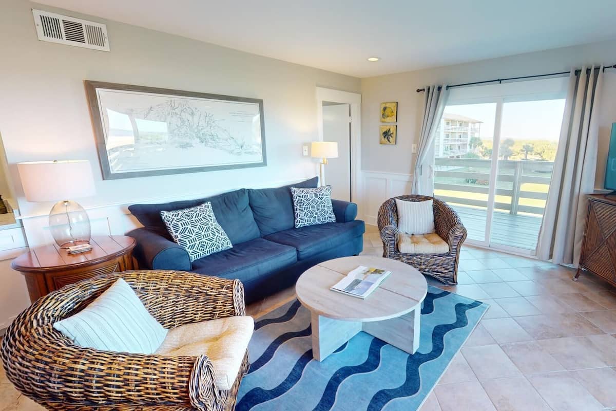 A modern living room with a sofa, two armchairs, a coffee table, a TV, and a large window with a view of buildings.
