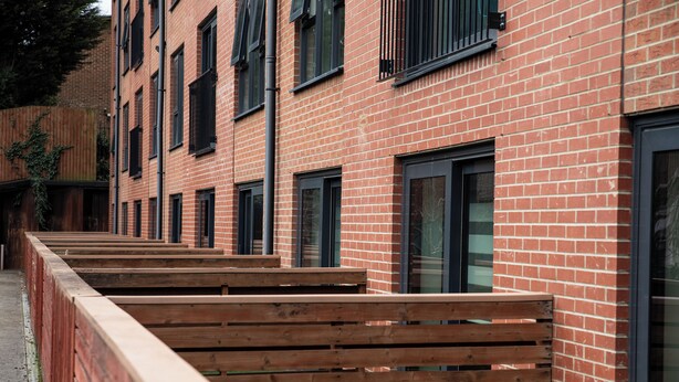 A row of wooden benches aligned with a brick building featuring multiple windows and a metal railing. - Student Only Tatton House  (Manchester)