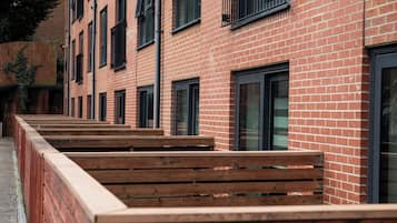 A row of wooden benches aligned with a brick building featuring multiple windows and a metal railing.