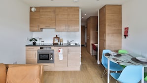 A modern kitchen with wooden cabinets, a built-in oven, and a dining area with blue chairs and a glass table. - Student Only Tatton House  (Manchester)