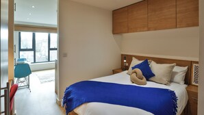 A modern bedroom with a large bed, a wooden headboard, and a view of the outside through large windows. - Student Only Tatton House  (Manchester)
