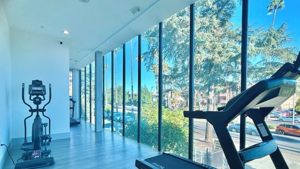 A gym with a treadmill and elliptical machine, large windows with a view of trees and a street. - Charming Glass-Front One Bedroom Spacious Living Free Parking (Los Angeles)