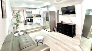 A modern kitchen with white cabinets, a microwave, and a refrigerator. There is a dining area with a gray sofa and a small round table. A flat-screen TV is mounted on the wall. - Charming Glass-Front One Bedroom Spacious Living Free Parking (Los Angeles)