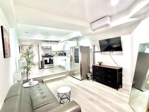 Eine moderne Küche mit weißen Schränken, einer Mikrowelle und einem Kühlschrank. Es gibt einen Essbereich mit einem grauen Sofa und einem kleinen runden Tisch. Ein Flachbildfernseher ist an der Wand montiert.