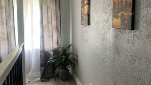 A room with a staircase, a potted plant, patterned curtains, and two wall-mounted paintings.