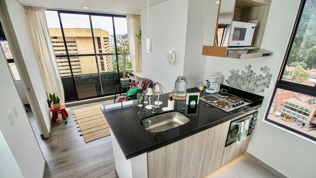 A modern kitchen with a black countertop, stainless steel sink, and a microwave. There is a window with a view of buildings and a red chair in the background. - Downtown Bogotá | Work & Stay (Bogotá)