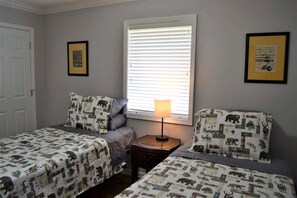 A bedroom with two beds, a nightstand, a window with blinds, and framed pictures on the walls.