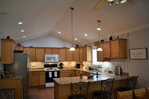 A kitchen with wooden cabinets, a stainless steel refrigerator, a microwave, an oven, and a sink. There are hanging lights, a bar area with stools, and a framed sign on the wall.