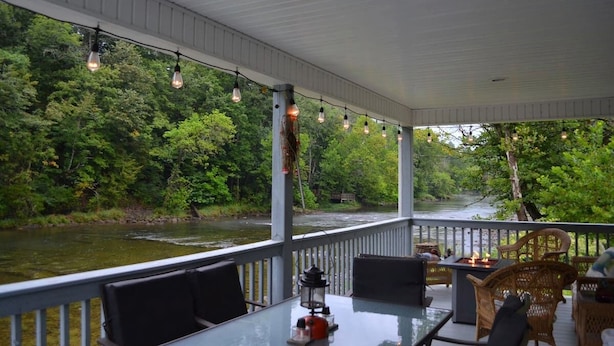 A covered outdoor deck with a glass table, wicker chairs, and a view of a river and forest. - Riverview Retreat Pad #5 (Bristol)