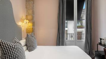 Habitación de hotel con una cama, cortinas a cuadros, una pared de piedra y un balcón con plantas en macetas.