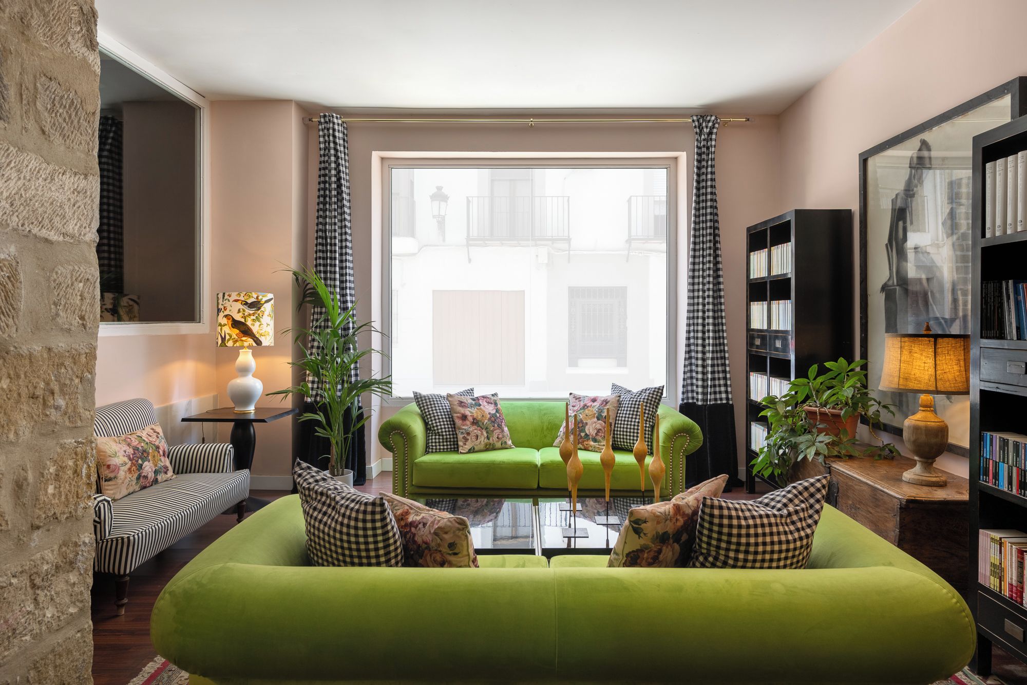 A living room with a green sofa, checkered pillows, a bookshelf, and a window with curtains.