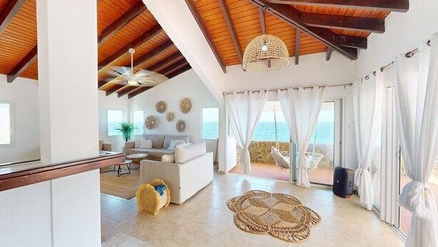 Amplia sala de estar con techos altos, ventilador de techo y vista al mar a través de grandes ventanales.