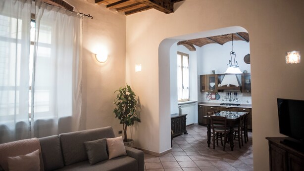 A living room with a sofa, a potted plant, and a kitchen area visible through an archway.