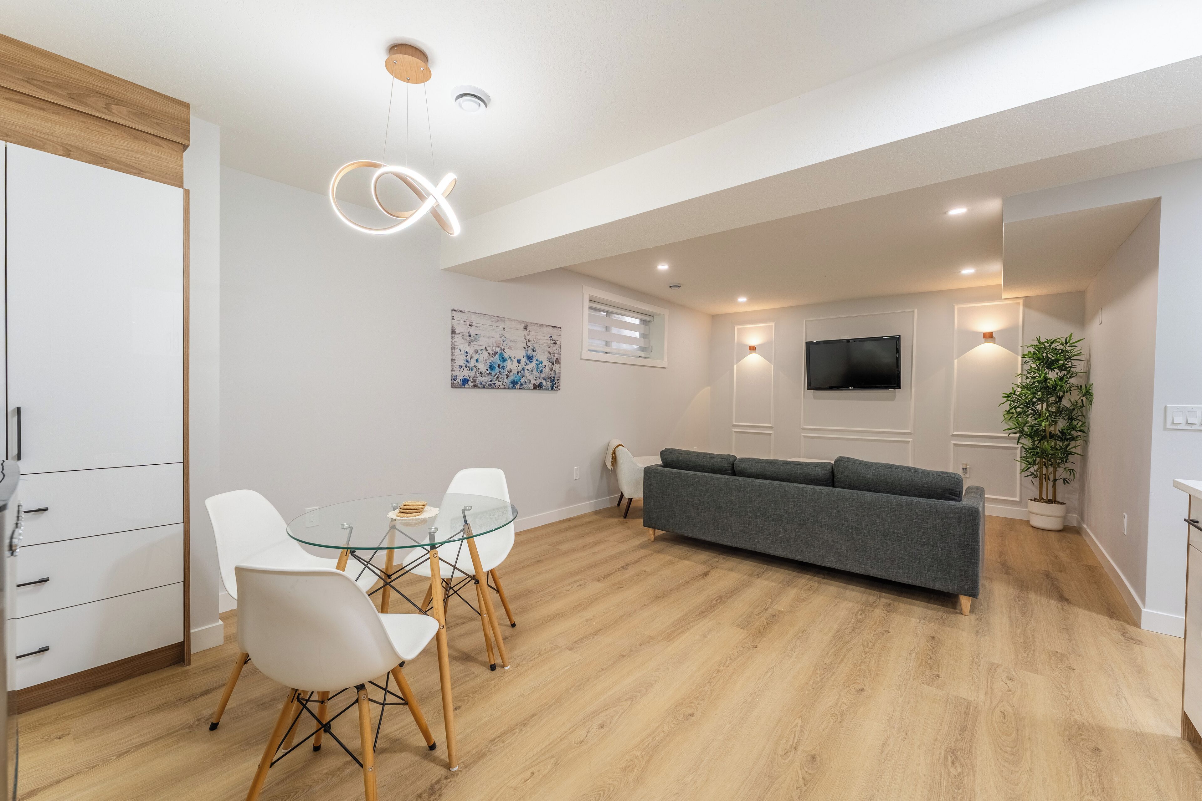 A modern living room with a grey sofa, a glass dining table with white chairs, a wooden floor, recessed lighting, a flat-screen TV, and a potted plant.