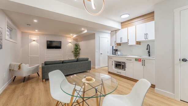 A modern kitchen with white cabinets, a microwave, and a sink. There is a dining area with a glass table and white chairs. A gray sofa is in the living area.