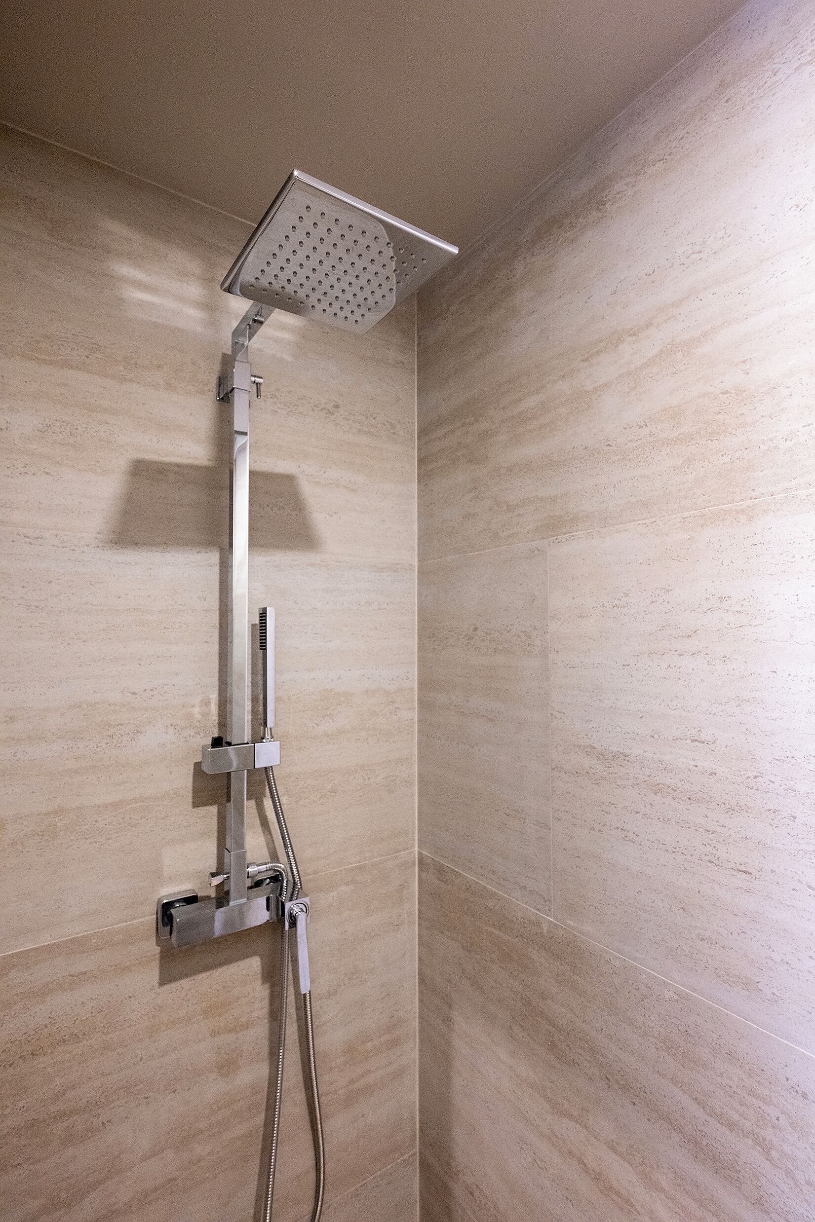 Une douche équipée d’un pommeau de douche à main et d’un pommeau de douche fixe au plafond, le tout installé sur un mur recouvert de carreaux de couleur beige clair.
