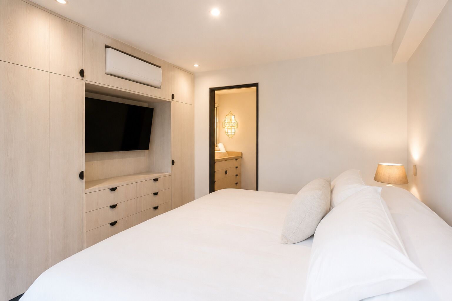 A bedroom with a bed, a TV mounted on a wooden cabinet, a bedside table with a lamp, and a wall-mounted light fixture.